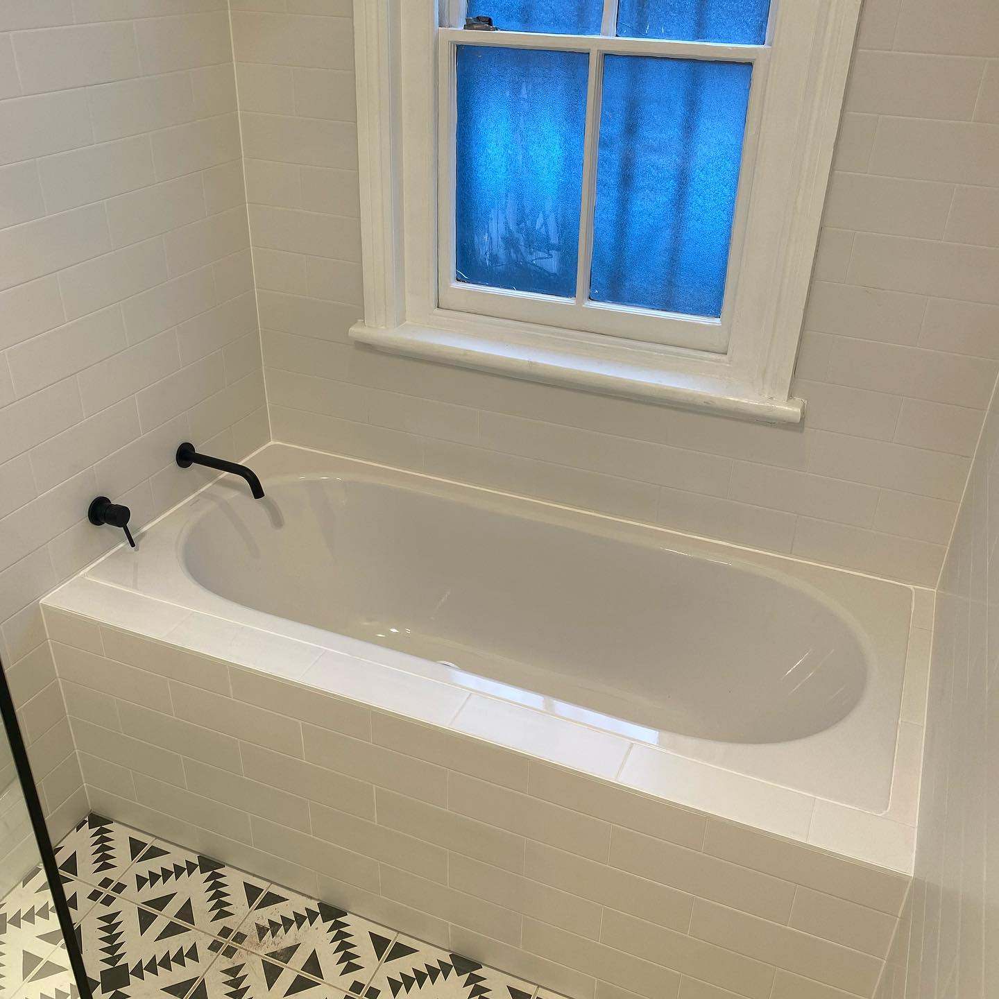 Built-in bath under a window with a black wall-mounted tap and patterned floor tile