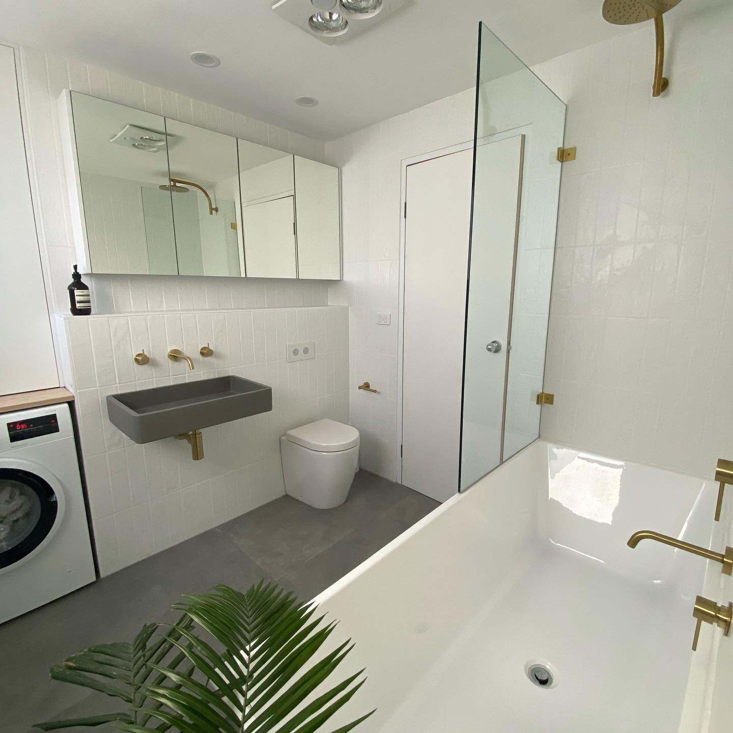 White bathroom and laundry with brass tapware, glass shower screen and a wall-hung basin